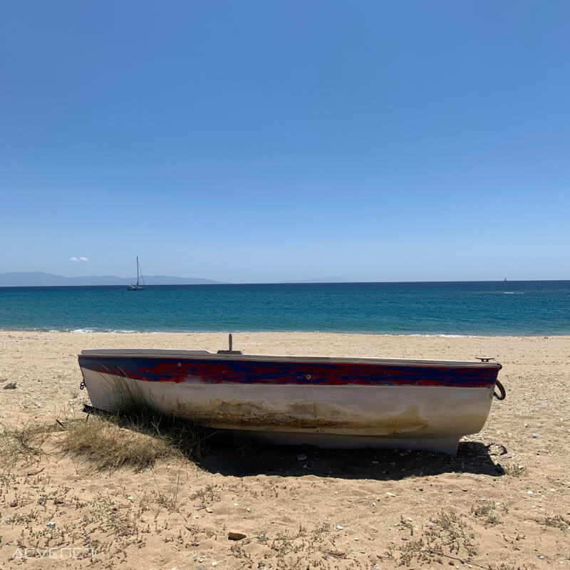 Petite embarcation sur la plage, mer et voilier en arrière-plan.