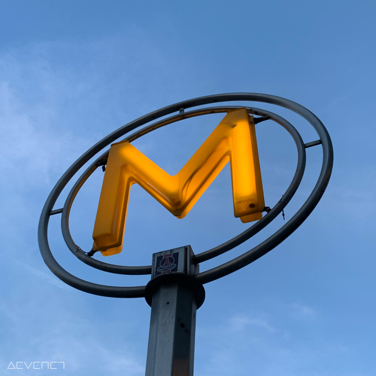 Panneau métro en forme M lumineux jaune encerclé par cercle métallique, avec le ciel bleu en fond.