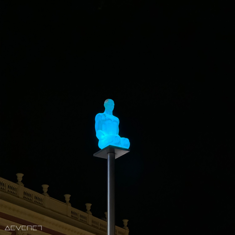 Dans une nuit noire, statue en résine éclairée en bleu représentant une personne en position accroupie.